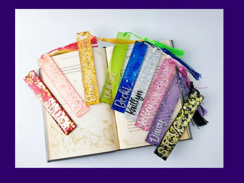Nine personalised glitter resin bookmarks in assorted colours arranged around an open book, each with a matching tassel