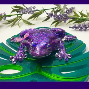 “Front-facing purple glitter resin frog with a butterfly on its back, sitting on a green acrylic monstera leaf.
