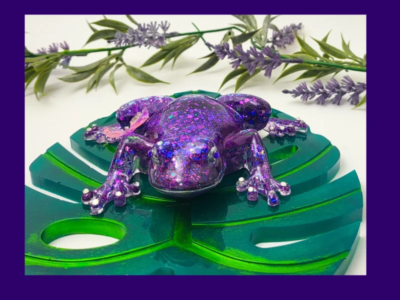 2. Kirnix Listing Primary โFront-facing purple glitter resin frog with a butterfly on its back, sitting on a green acrylic monstera leaf.