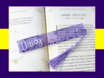 Violet glitter bookmark personalised with the name Daisy, lying across an open book with a light purple tassel.