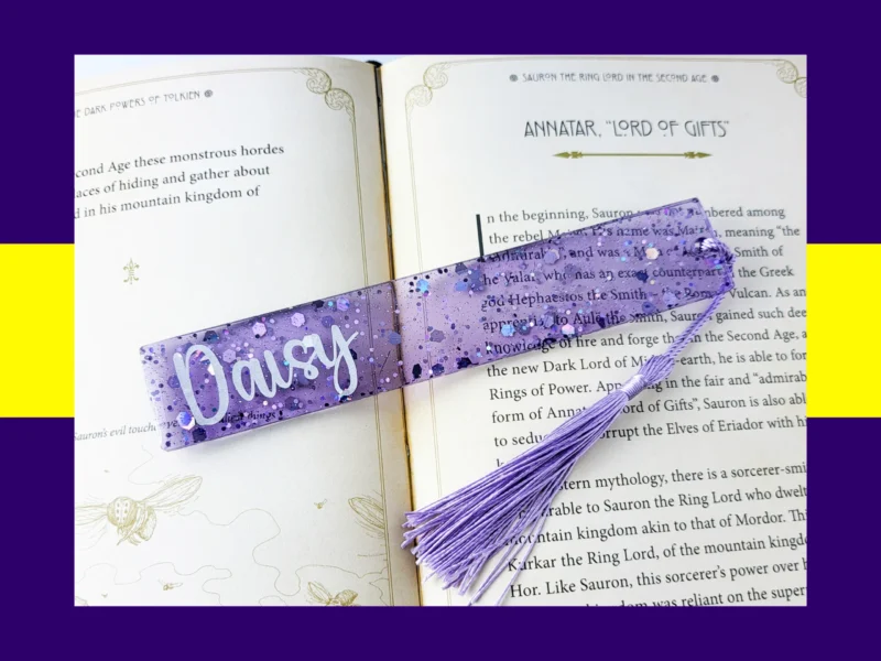 Violet glitter bookmark personalised with the name Daisy, lying across an open book with a light purple tassel.