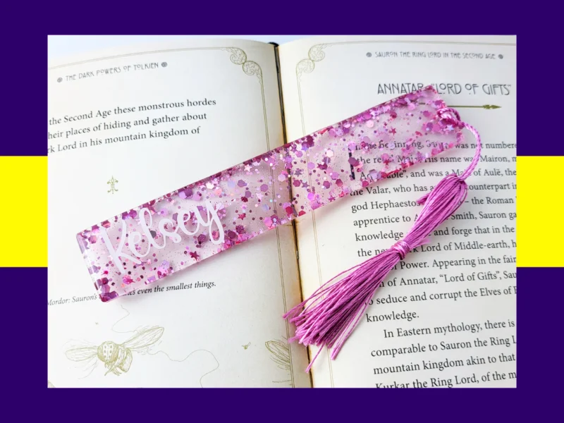 Pink glitter bookmark personalised with the name Daisy, shown across the pages of an open book with a matching pink tassel.
