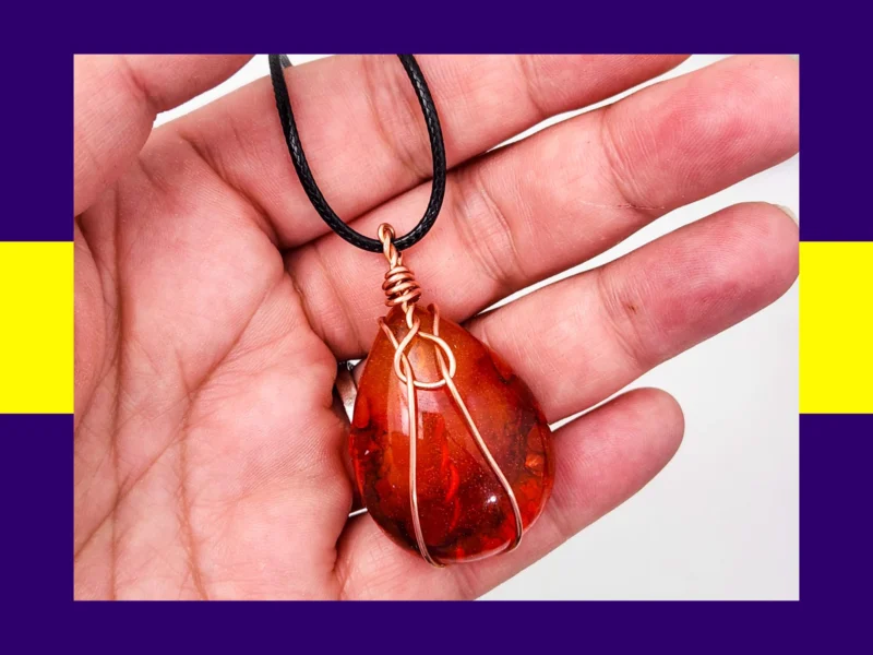 Red and amber autumn glow resin teardrop pendant with copper wire wrap shown in hand for scale – handmade necklace by Kirnix Creations