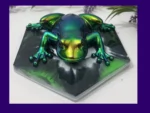 Chrome colour-shift resin frog in green, gold and teal tones, displayed on a black hexagonal base with foliage behind