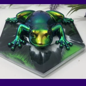 Chrome colour-shift resin frog in green, gold and teal tones, displayed on a black hexagonal base with foliage behind