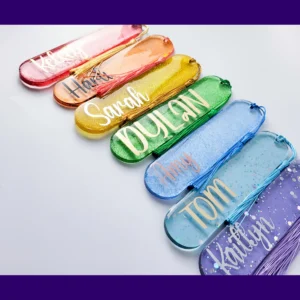 Oval glitter resin bookmarks in a rainbow of colours with personalised names and tassels arranged in a row.