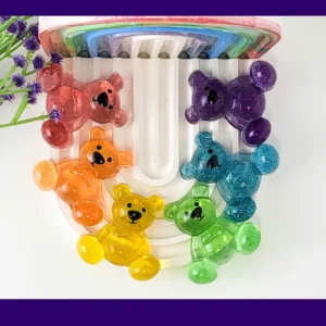 Six glitter resin Worry Bears in rainbow colours arranged on arch tray with purple flowers, colourful anxiety calming gifts