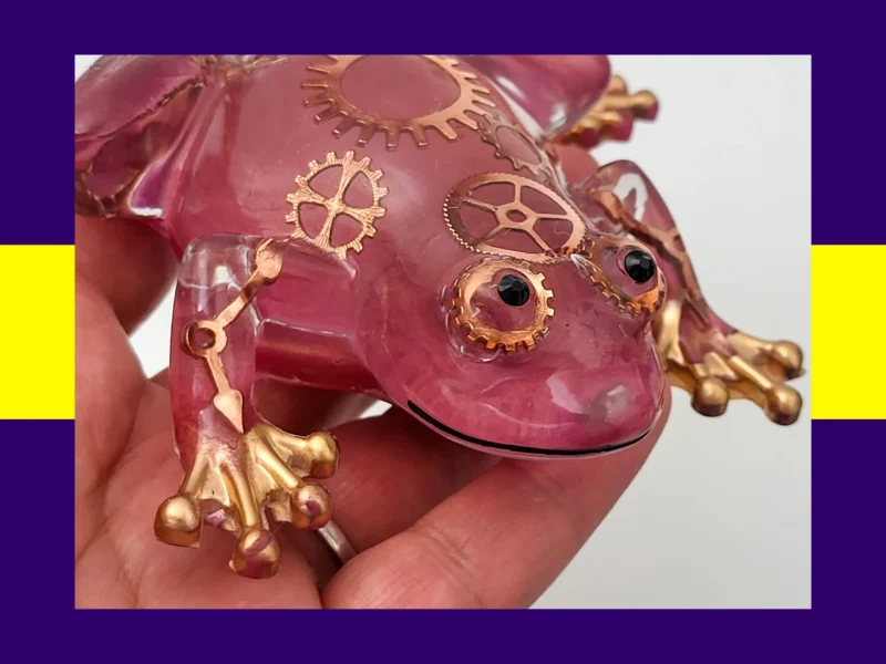 Hand holding rose-pink steampunk resin frog showing detailed gears and glossy finish.