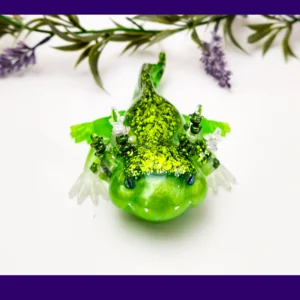Front-facing glitter green resin axolotl surrounded by decorative foliage.