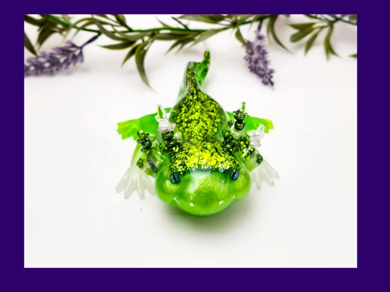 Front-facing glitter green resin axolotl surrounded by decorative foliage.