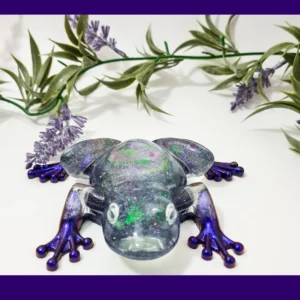 Glittery resin frog ornament with metallic purple legs and a smoky “galaxy” body, photographed from the front on a white background with decorative greenery.