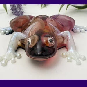 Front-on photo of the Kirnix Creations Bloom Frog, a glossy handmade resin frog with marbled burgundy, plum and amber body, pearly white legs and googly eyes, framed by a purple border.