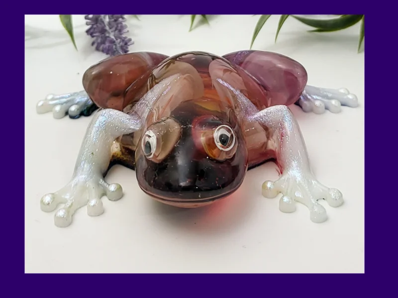 KC-FRG-BLOOM-OOAK-001002 Front-on photo of the Kirnix Creations Bloom Frog, a glossy handmade resin frog with marbled burgundy, plum and amber body, pearly white legs and googly eyes, framed by a purple border.