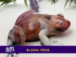 KC-FRG-BLOOM-OOAK-001003 Side view of the Bloom Frog resin sculpture with a marbled burgundy/amber body and pearly legs; a purple banner along the bottom reads โBLOOM FROGโ with a small KN logo.