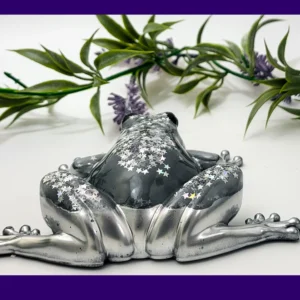 Rear view of a silver and charcoal resin frog with silver star glitter across its back, photographed on a white background with greenery.