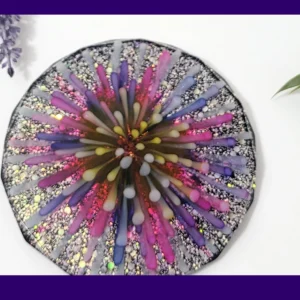 Top-down photo of a handmade resin coaster with a “firework burst” design in pink, purple, yellow and white over a silver glitter background, framed with a purple border.