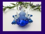 KNC-AXO-ICE-00101_Hero Fire & Ice axolotl (ice version) in blue glitter resin with translucent frills, photographed on a white background with greenery, framed by a purple border.