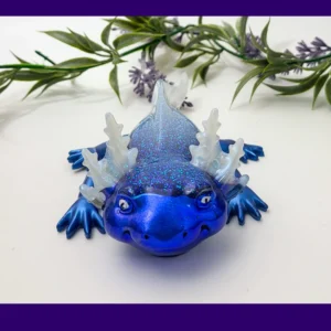 Fire & Ice axolotl (ice version) in blue glitter resin with translucent frills, photographed on a white background with greenery, framed by a purple border.