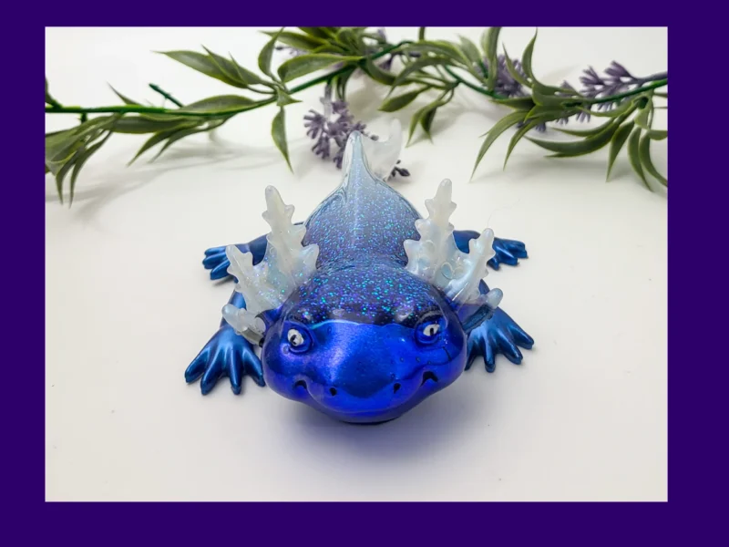 KNC-AXO-ICE-00101_Hero Fire & Ice axolotl (ice version) in blue glitter resin with translucent frills, photographed on a white background with greenery, framed by a purple border.