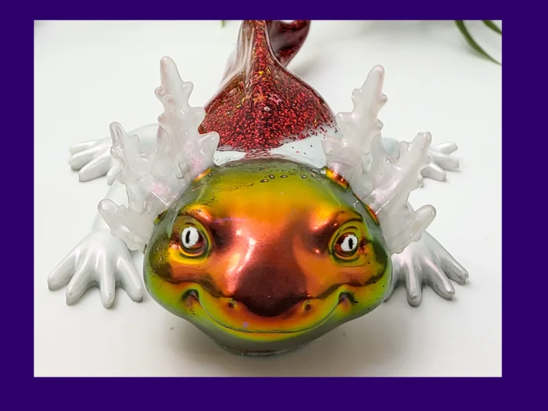 Fire axolotl with iridescent gold-green face and red glitter back on a white background with greenery, framed by a purple border.