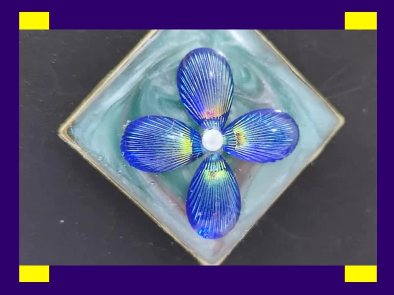 Close-up of a square resin pendant with iridescent blue petal design inside a metal frame.