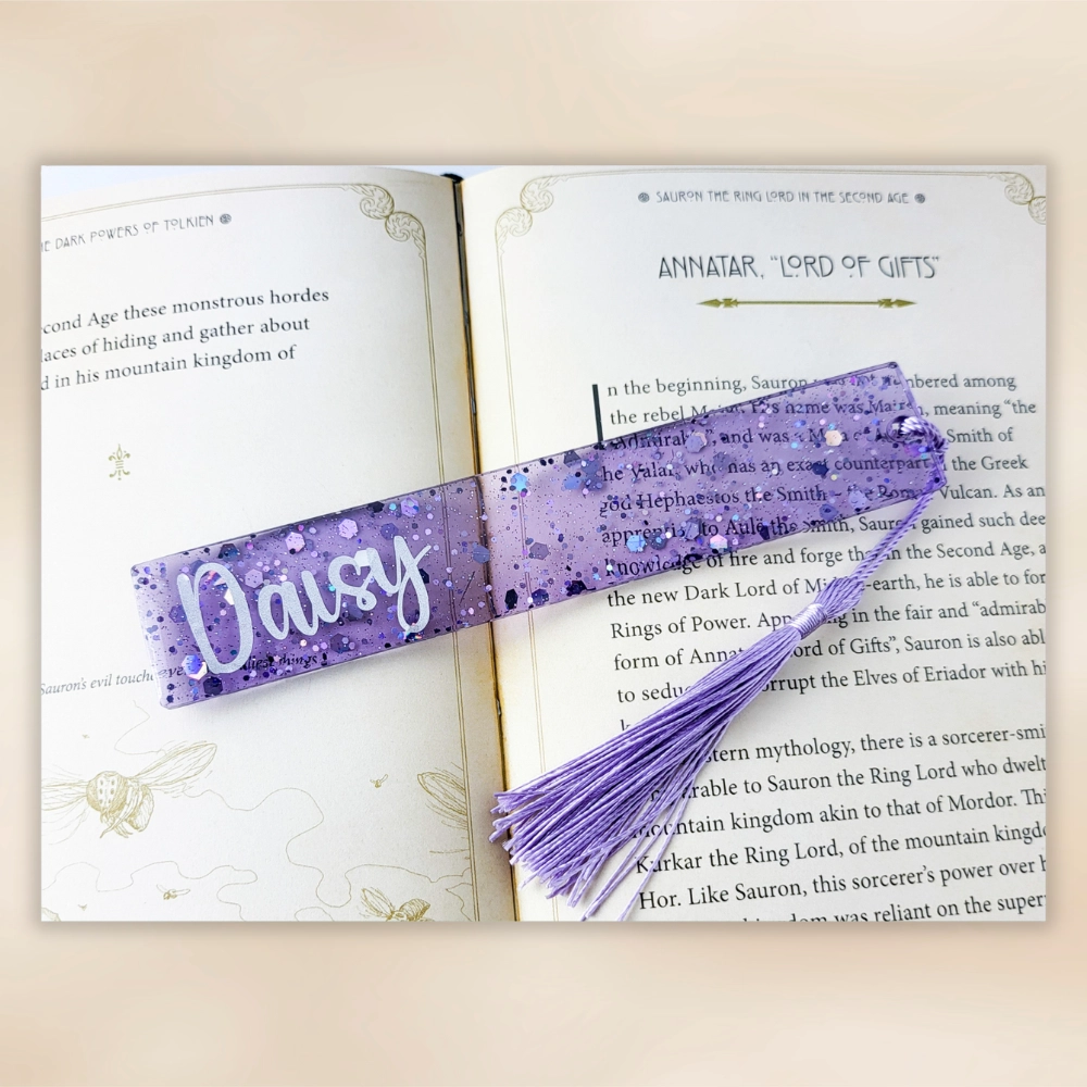 Purple personalised resin bookmark with tassel resting across an open book on a warm neutral background.