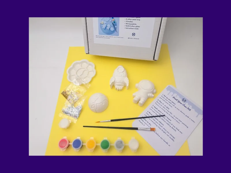 Paint your own space kit laid out on yellow background with paints brushes and instructions