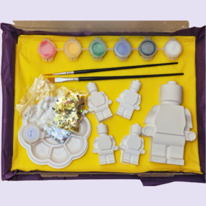 Open yellow gift box with craft supplies: pastel paint pots, two brushes, white plastic shapes, sequins, and a round palette.