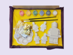 Open yellow gift box with craft supplies: pastel paint pots, two brushes, white plastic shapes, sequins, and a round palette.