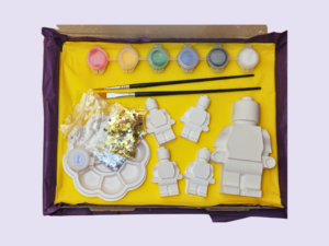 Open yellow gift box with craft supplies: pastel paint pots, two brushes, white plastic shapes, sequins, and a round palette.
