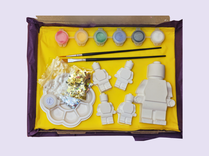 Open yellow gift box with craft supplies: pastel paint pots, two brushes, white plastic shapes, sequins, and a round palette.