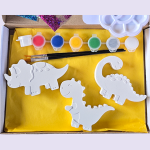Craft kit with six paint pots (red, blue, yellow, green, gray/white) and white dinosaur-shaped cutouts on a yellow mat, plus brushes in a box.