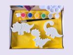 Craft kit with six paint pots (red, blue, yellow, green, gray/white) and white dinosaur-shaped cutouts on a yellow mat, plus brushes in a box.