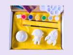 KNP-PYO-SPACE_Hero photo Open craft kit showing white space-themed shapes (astronaut and rocket) laid on yellow tissue with small paint pots in pink, blue, orange, green, gray, and white inside a box.