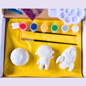 Open craft kit showing white space-themed shapes (astronaut and rocket) laid on yellow tissue with small paint pots in pink, blue, orange, green, gray, and white inside a box.