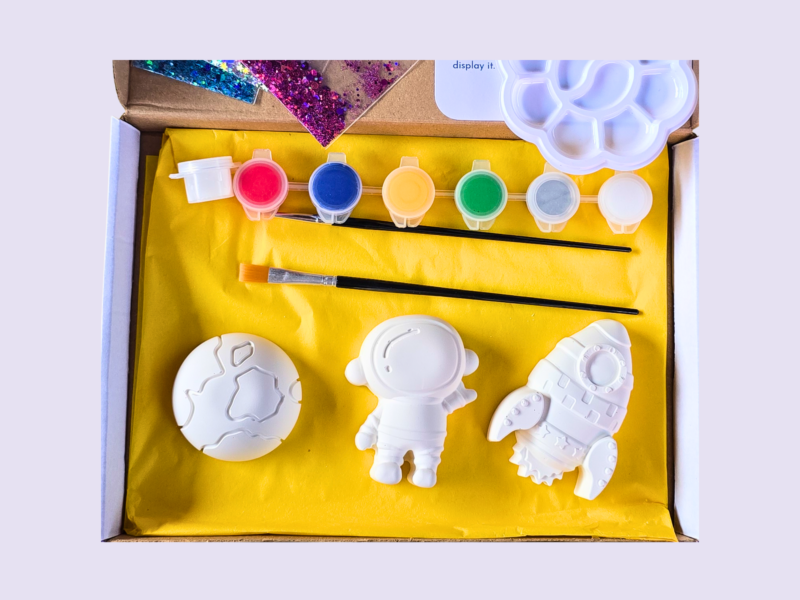 KNP-PYO-SPACE_Hero photo Open craft kit showing white space-themed shapes (astronaut and rocket) laid on yellow tissue with small paint pots in pink, blue, orange, green, gray, and white inside a box.