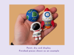 KNP-PYO-SPACE_Painted result Hand holding a small blue Earth-like ball and a white and red rocket figurine painted in bright colors, against a peach background; suggests a space-themed craft project.