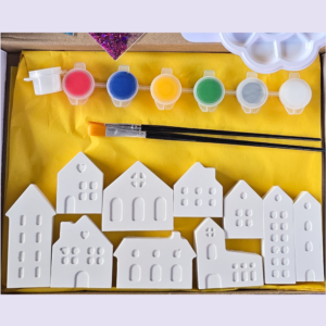 Row of white plastic house shapes with window cutouts laid on a yellow mat, above them are small paint pots in pink, blue, yellow, green, gray, and brushes.
