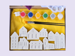 Row of white plastic house shapes with window cutouts laid on a yellow mat, above them are small paint pots in pink, blue, yellow, green, gray, and brushes.