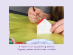 Child’s hand paints a small white ceramic house with a red roof using a fine brush in a craft project.