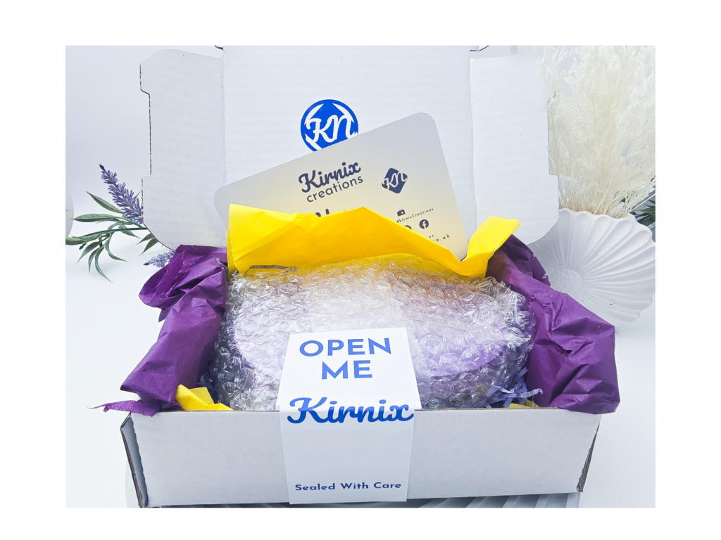 Kirnix eco-friendly packaging with a handmade resin order wrapped in protective bubble wrap, purple and yellow tissue paper, and branded studio inserts inside a white postal box.