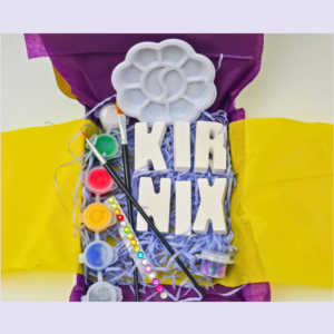 Colorful craft supplies on a purple and yellow backdrop, including a white flower-shaped paint palette, brushes, and paint pots.
