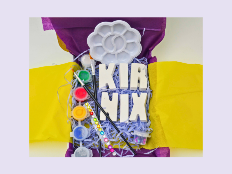 Colorful craft supplies on a purple and yellow backdrop, including a white flower-shaped paint palette, brushes, and paint pots.