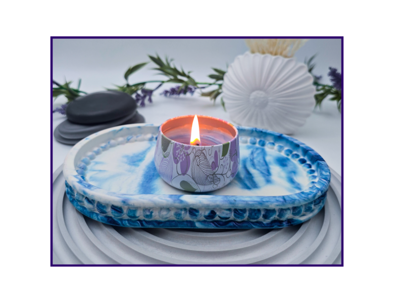 Lit candle in a patterned holder resting on a blue marbled plate with lavender and smooth stones in the background for decoration.