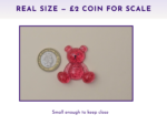 Small pink glitter bear figurine beside a £2 coin on a light surface to show scale.