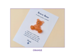 Orange glitter bear charm placed on a white card that reads 'Brave Bear' and 'A little pocket comfort' against a pale beige background.