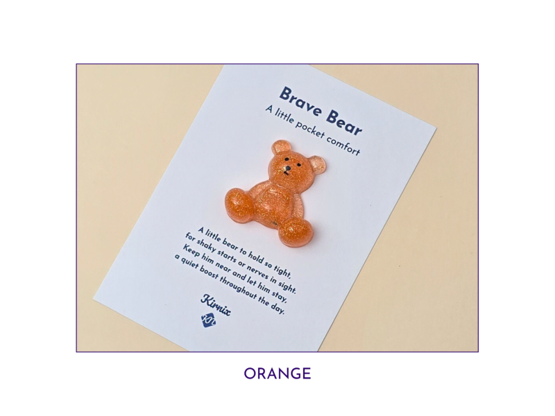Orange glitter bear charm placed on a white card that reads 'Brave Bear' and 'A little pocket comfort' against a pale beige background.