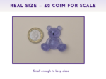 Purple glitter teddy bear figure beside a UK two-pence coin on a light surface, showing scale for size comparison.