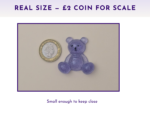 Purple glitter teddy bear figure beside a UK two-pence coin on a light surface, showing scale for size comparison.