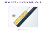 KXK-RBMK-07 Glittery gold rectangular pendant next to a black ruler for scale, with a UK one-pound coin nearby for reference.